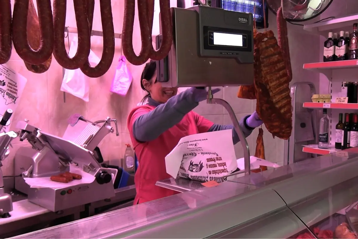 Mujer sonriente atendiendo en una charcutería mientras pesa embutidos detrás de un mostrador