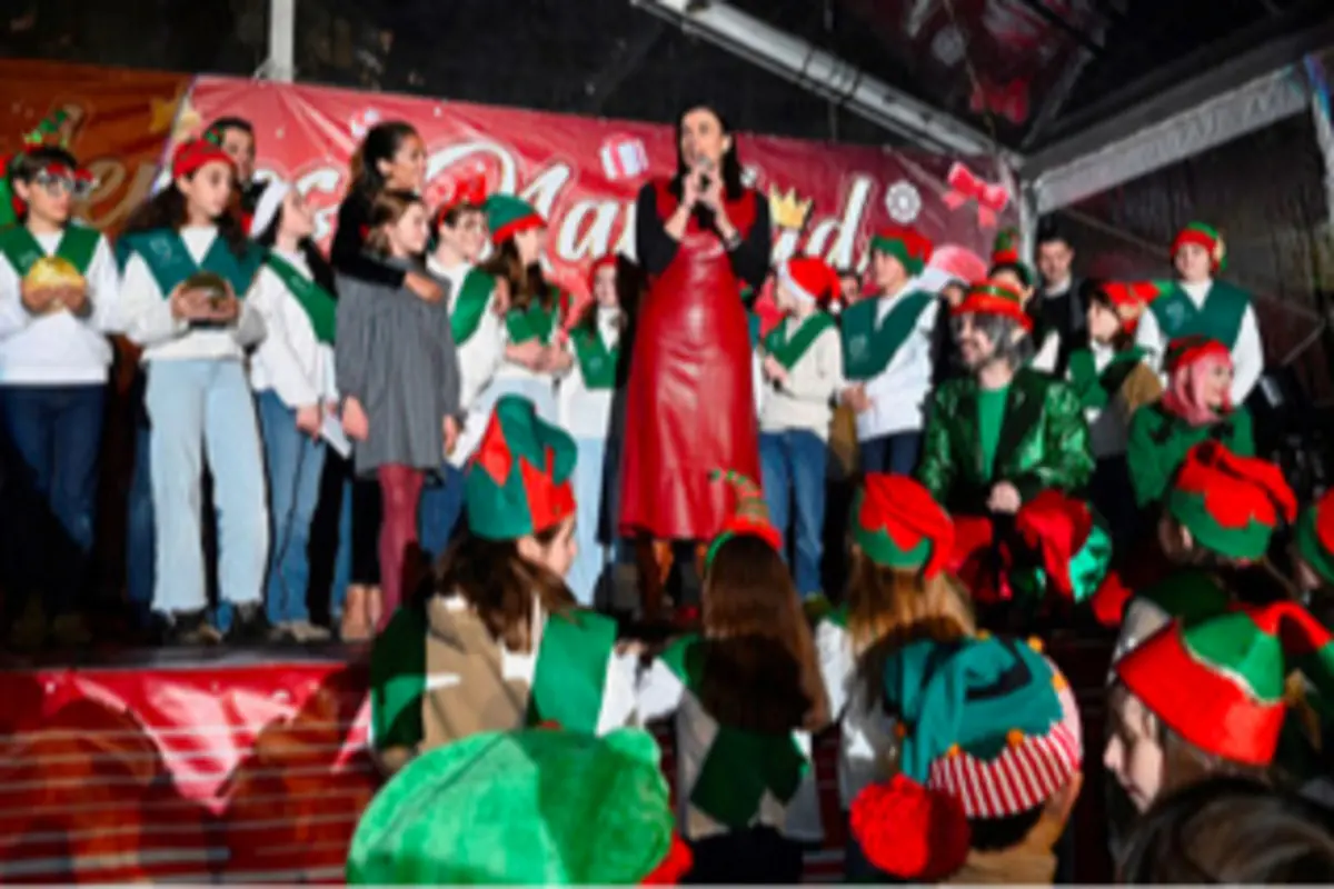 Grupo de niños y adultos disfrazados de elfos en un escenario festivo de Navidad mientras una mujer con vestido rojo habla al micrófono