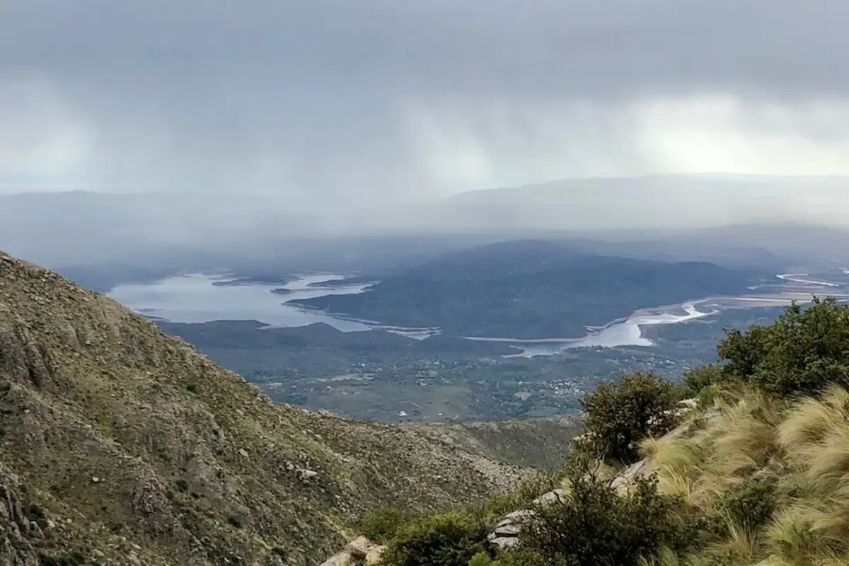 Paisaje de montañas con vegetación en primer plano y un lago serpenteante al fondo bajo un cielo nublado