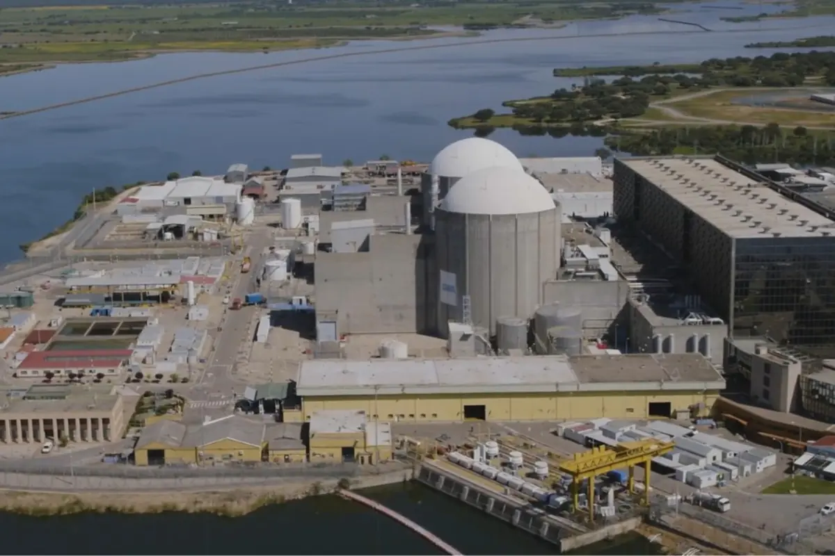 Vista aérea de una planta nuclear junto a un cuerpo de agua rodeada de edificios industriales y áreas verdes al fondo