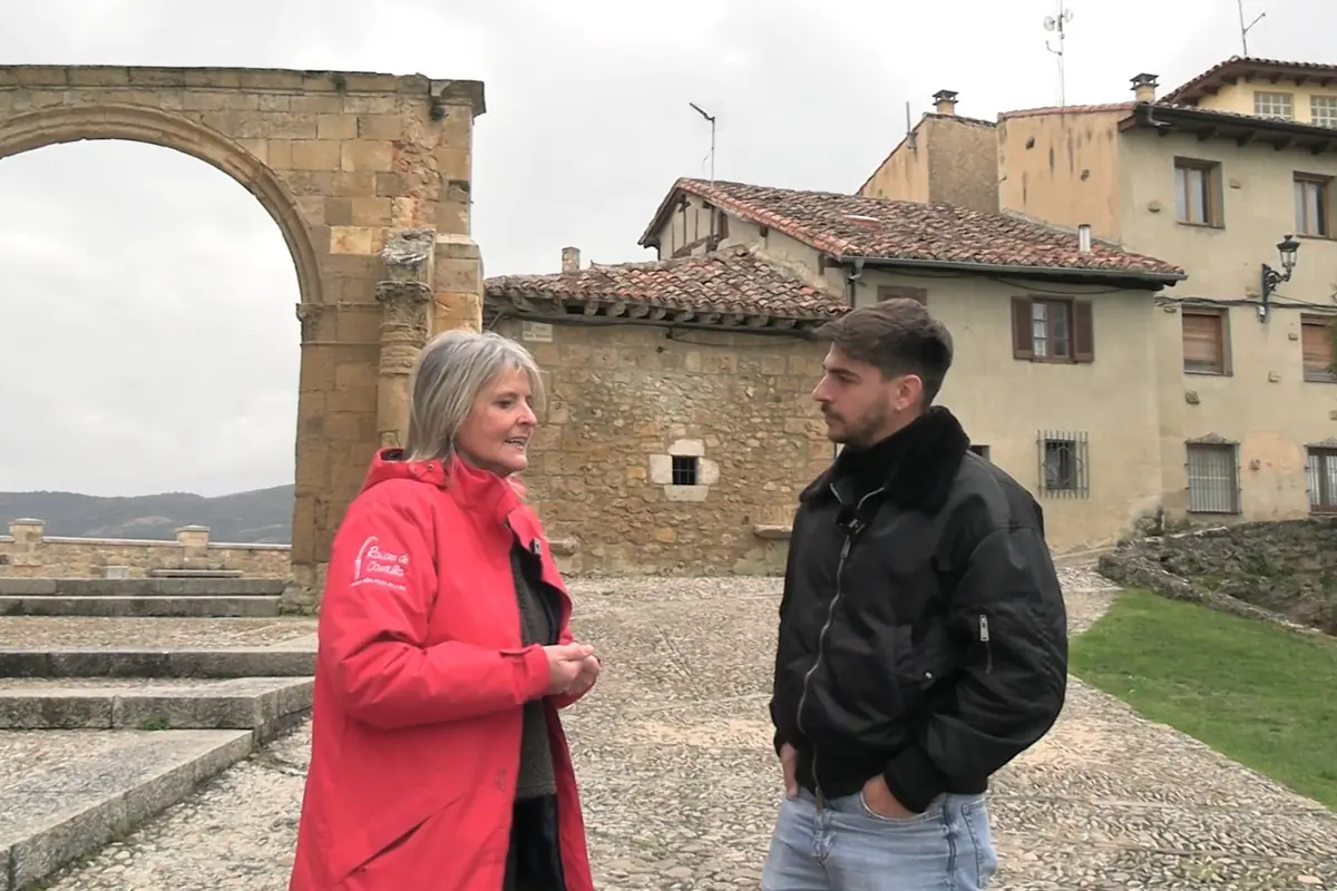 Dos personas conversan al aire libre frente a una construcción antigua de piedra y casas con tejados en un pueblo