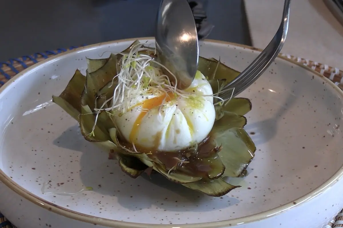Un huevo poché servido sobre hojas de alcachofa con brotes y salsa, siendo cortado con cuchara y tenedor en un plato blanco