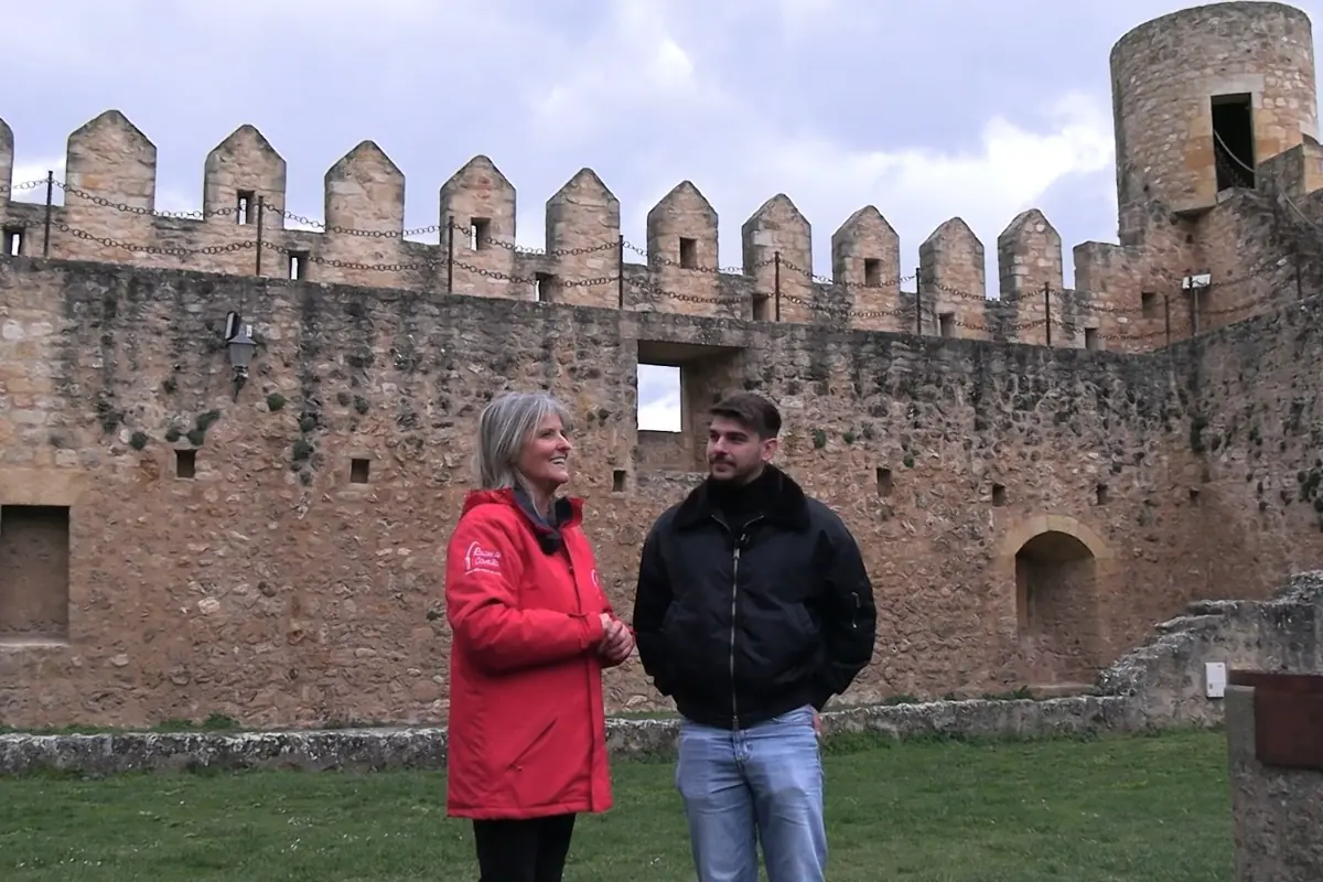 Dos personas conversan frente a una muralla de piedra antigua con almenas y una torre redonda, en un entorno al aire libre con césped.