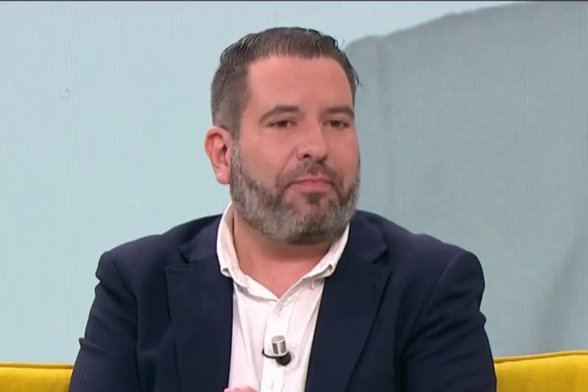 Hombre de mediana edad con barba canosa y traje oscuro sentado en un sofá amarillo durante una entrevista en televisión