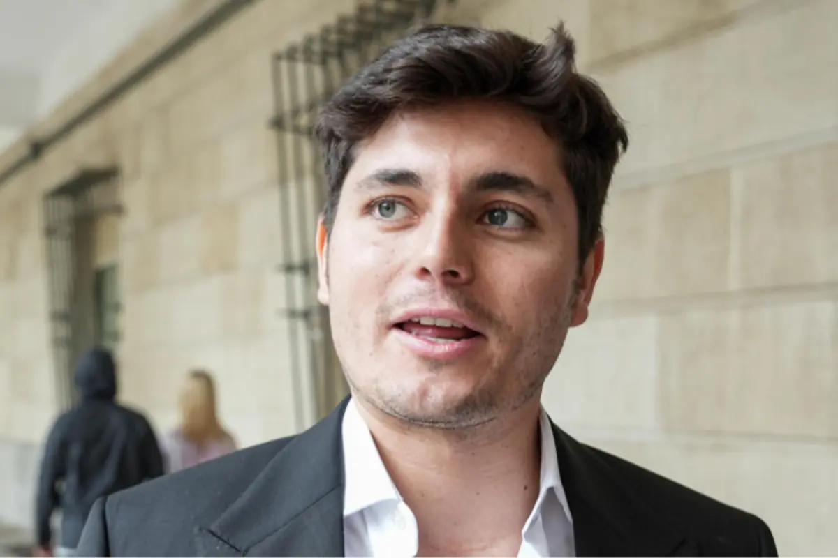 Joven de cabello corto y oscuro con saco negro y camisa blanca hablando al aire libre frente a una pared clara