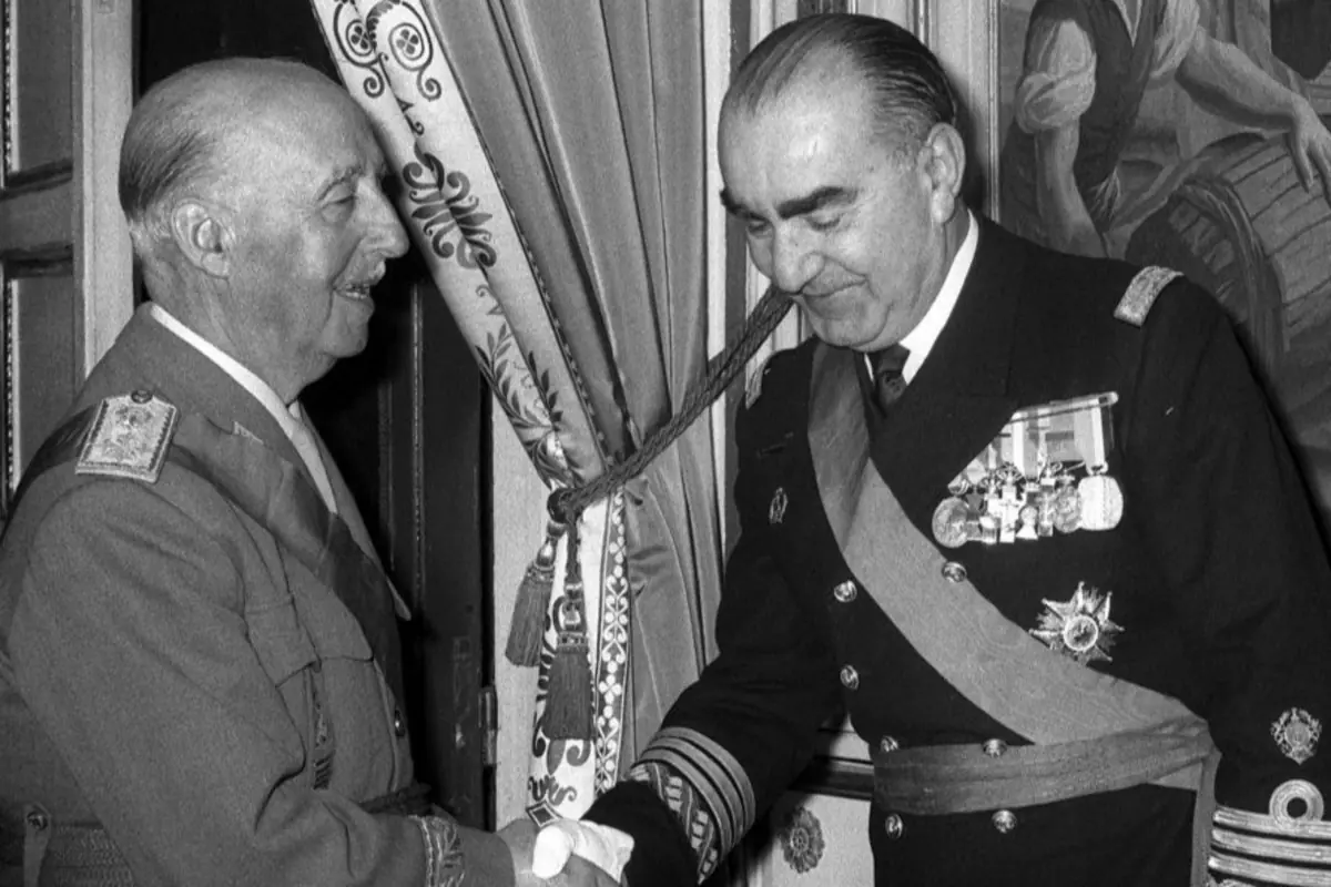 Dos hombres mayores con uniformes militares y condecoraciones se dan la mano en una sala con cortinas decoradas