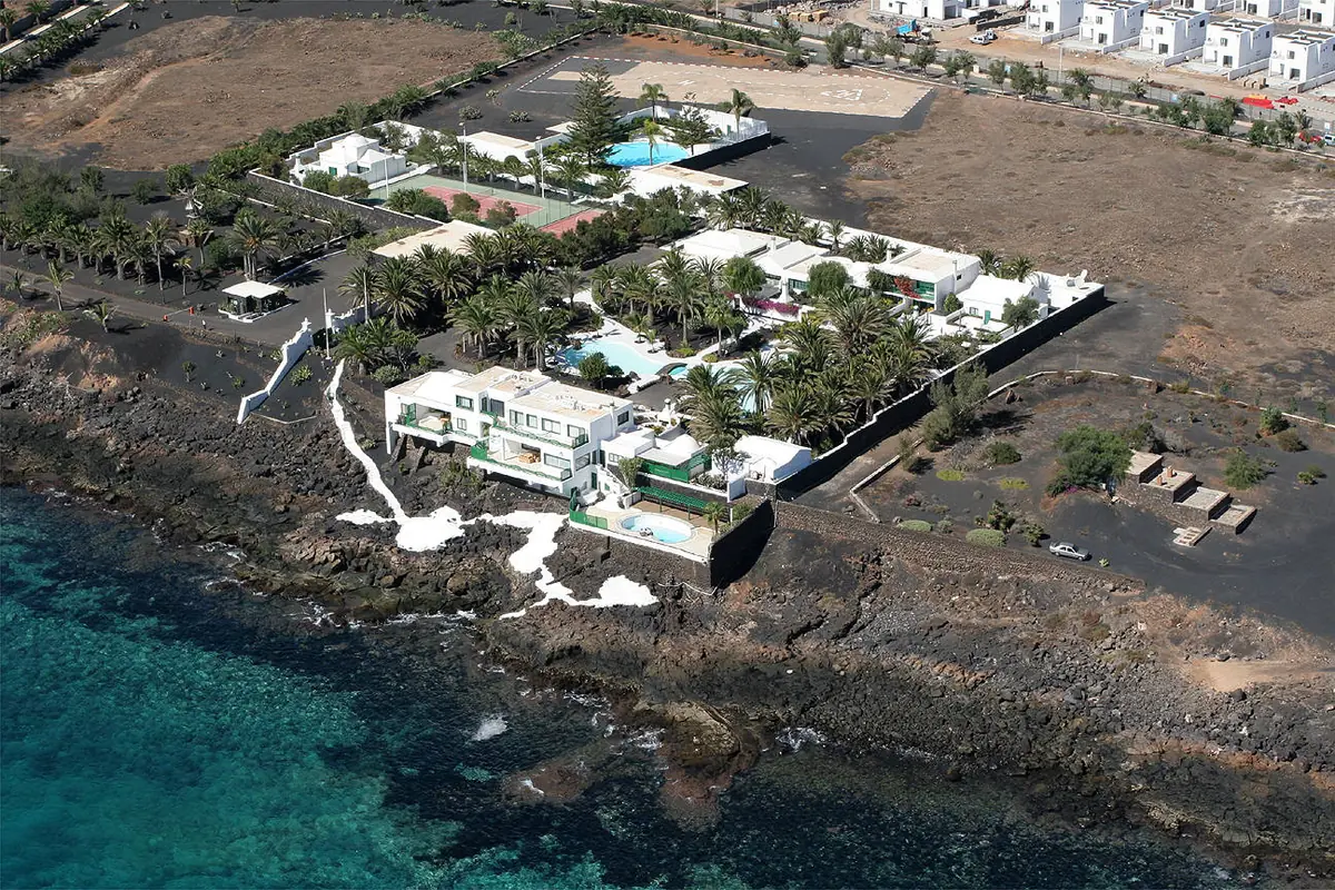 Vista aérea de una lujosa mansión blanca con piscina ubicada junto al mar, rodeada de palmeras y jardines, en una zona volcánica y rocosa