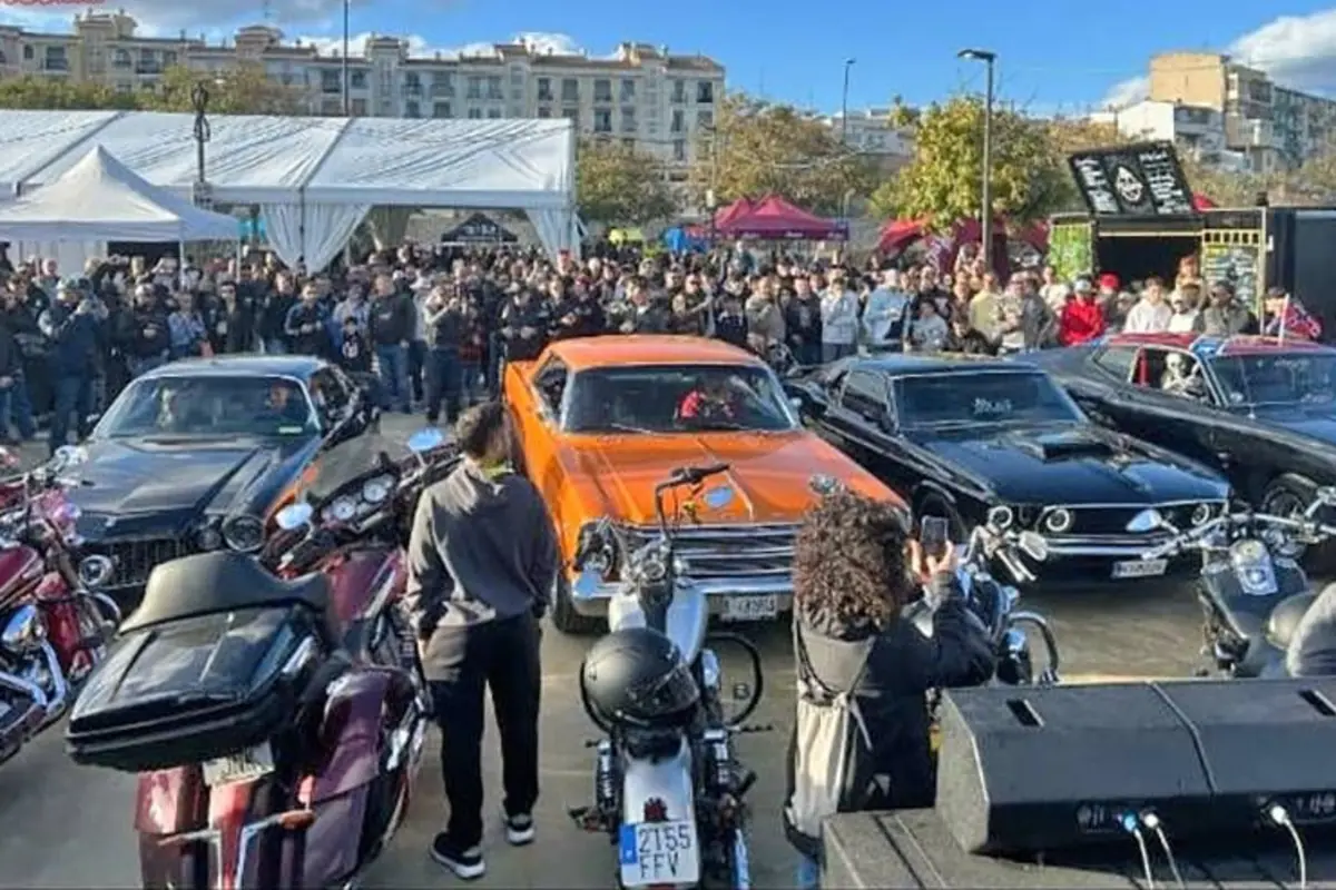 Evento al aire libre con una multitud observando autos clásicos y motocicletas estacionados, destacando un coche naranja en el centro