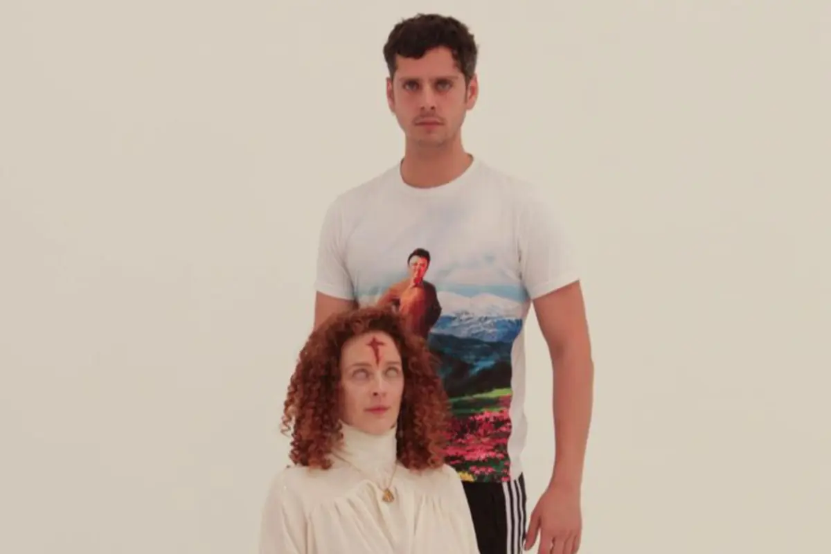 Un hombre de pie con camiseta blanca estampada y una mujer sentada con cabello rizado y una cruz roja en la frente sobre un fondo blanco