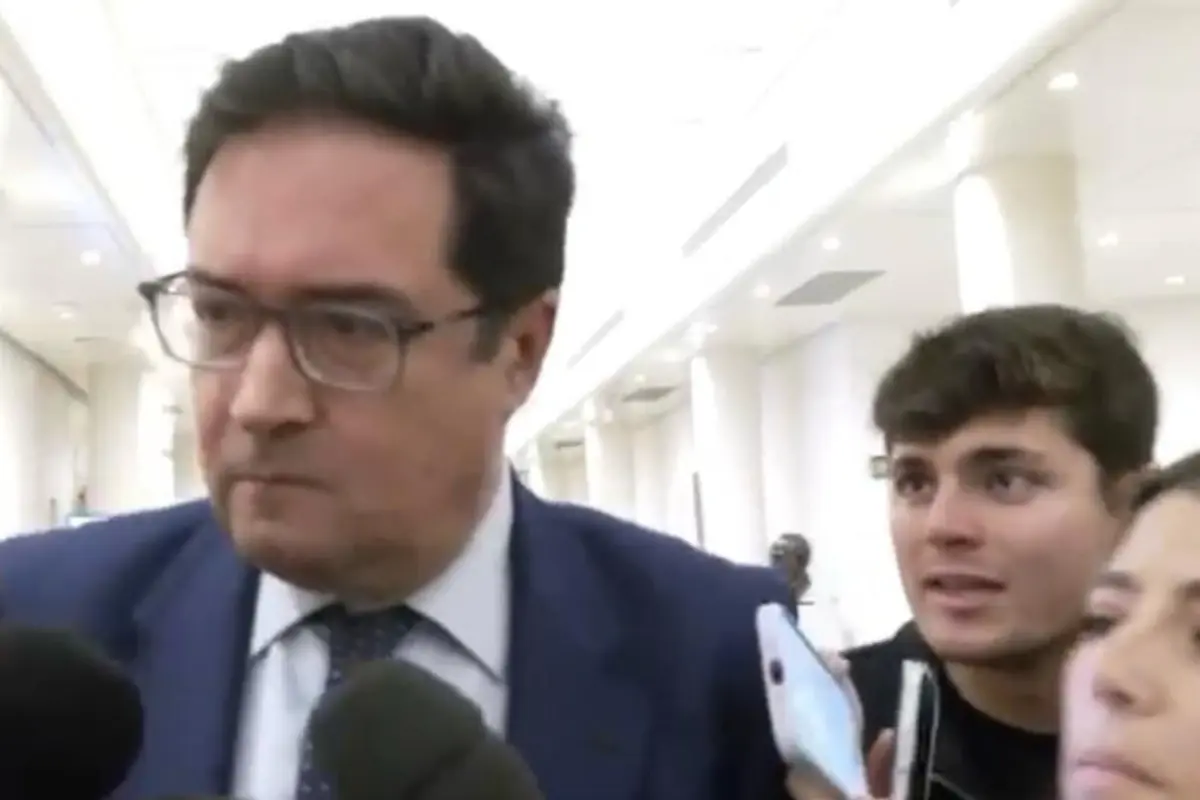 Hombre con traje azul y gafas camina por un pasillo mientras varios periodistas lo rodean con micrófonos y teléfonos móviles