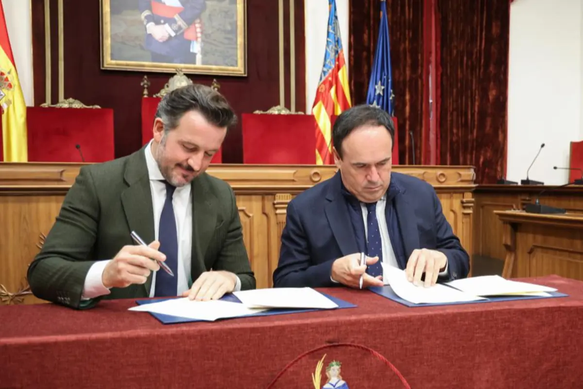 Dos hombres con traje firman documentos en una mesa cubierta con mantel rojo en un salón institucional con banderas y un retrato oficial al fondo