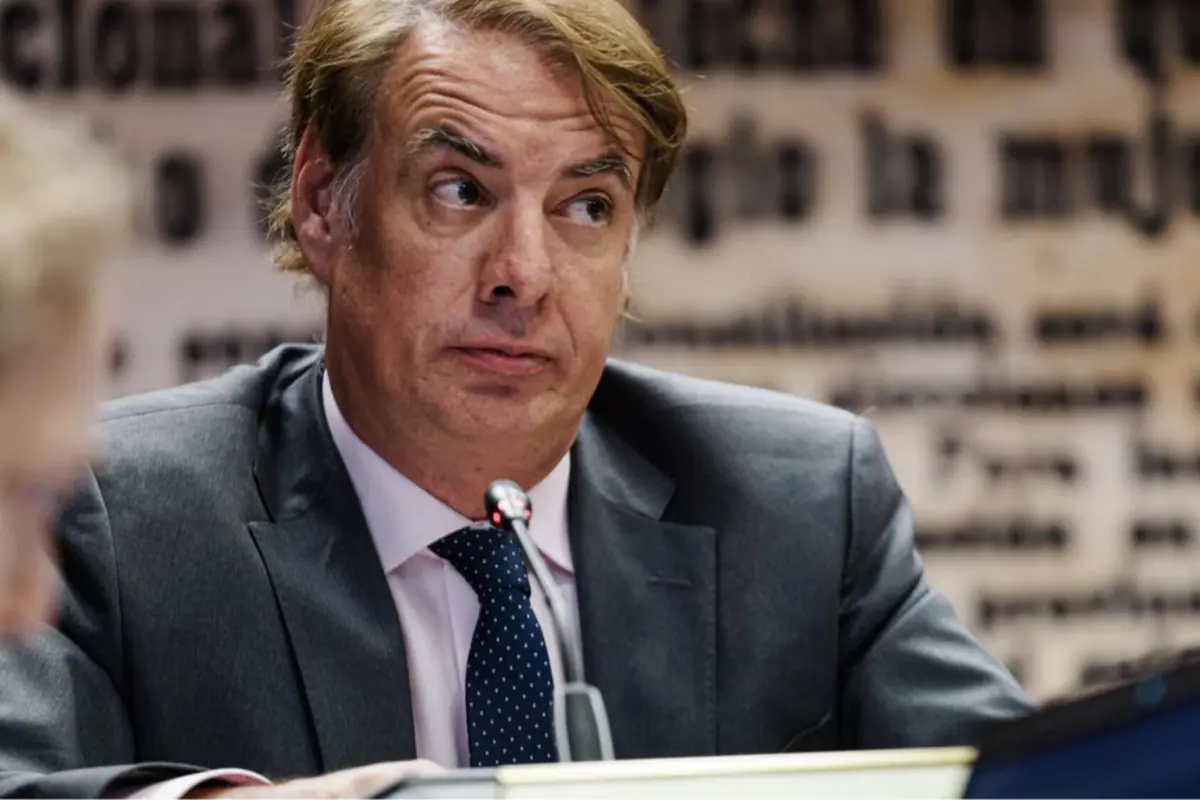 Hombre de traje y corbata azul con puntos blancos sentado frente a un micrófono mirando hacia un lado en un entorno formal