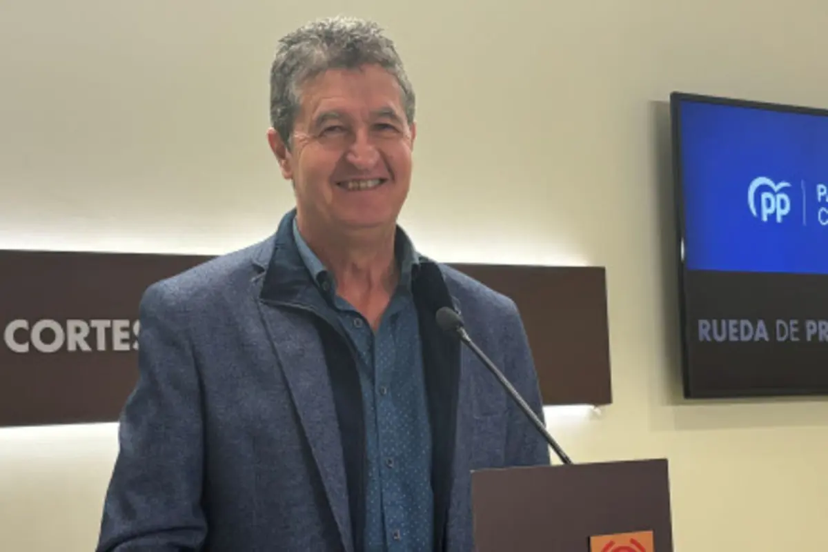 Hombre sonriente de cabello canoso hablando en una rueda de prensa frente a un micrófono con pantallas y carteles de fondo Hombre sonriente de cabello canoso hablando en una rueda de prensa frente a un micrófono con pantallas y carteles de fondo
