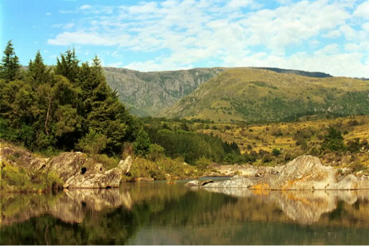 Paisaje de montaña con lago tranquilo en primer plano que refleja rocas y árboles bajo un cielo azul con nubes dispersas Paisaje de montaña con lago tranquilo en primer plano que refleja rocas y árboles bajo un cielo azul con nubes dispersas