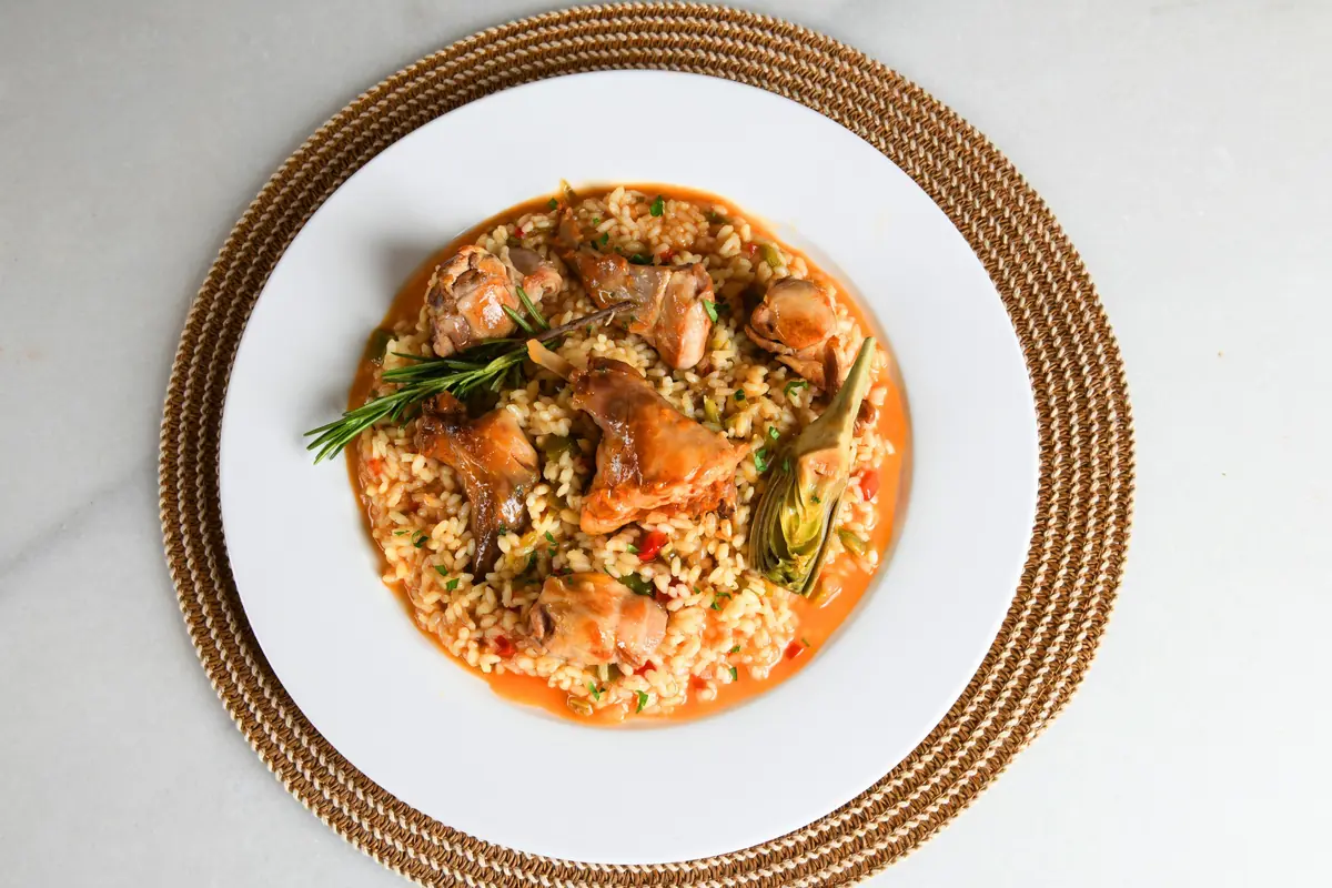 Plato de arroz meloso con trozos de conejo, alcachofa y una ramita de romero servido en un plato blanco sobre un mantel individual trenzado.