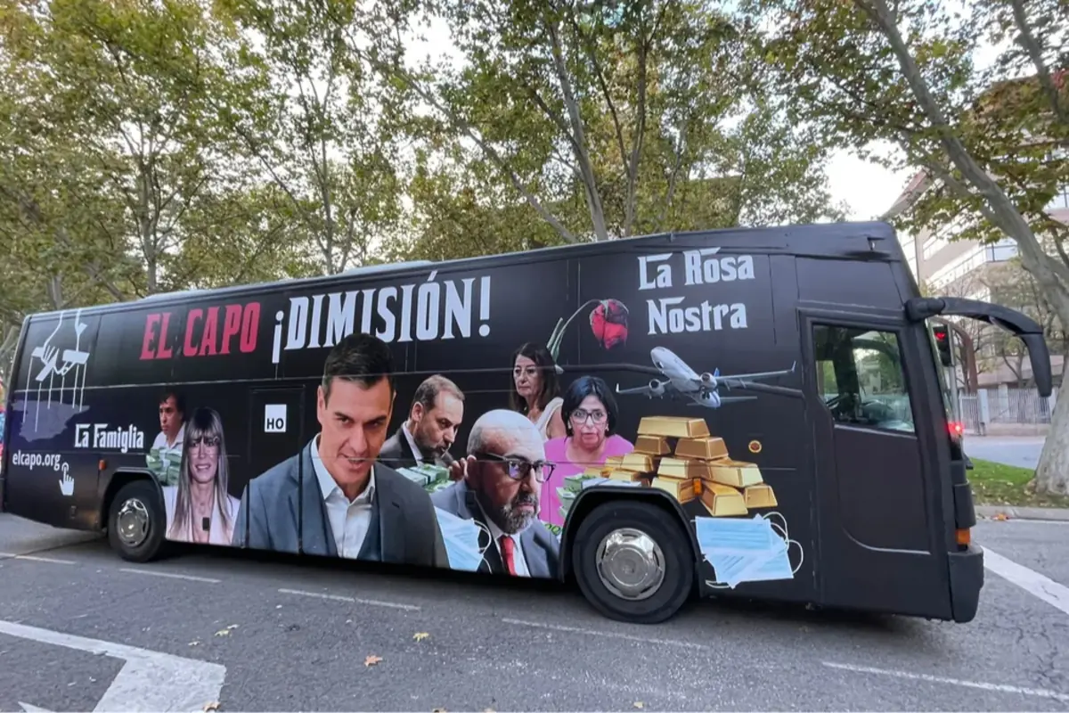 Autobús negro con imágenes de varios políticos, lingotes de oro, mascarillas, un avión y textos que dicen El Capo Dimisión La Rosa Nostra y La Famiglia, estacionado en una calle con árboles de fondo