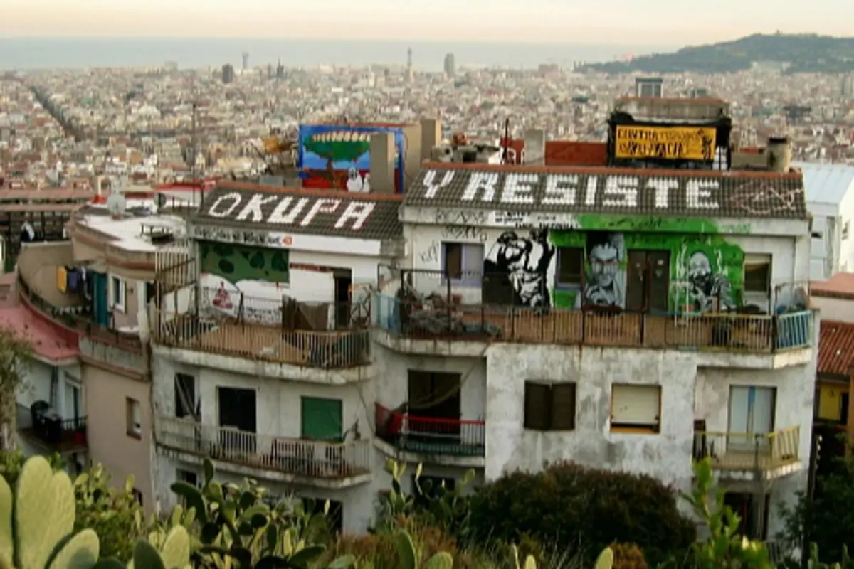 Edificio ocupado con fachadas llenas de grafitis y el mensaje OKUPA Y RESISTE en el tejado frente a una vista panorámica de la ciudad Edificio ocupado con fachadas llenas de grafitis y el mensaje OKUPA Y RESISTE en el tejado frente a una vista panorámica de la ciudad