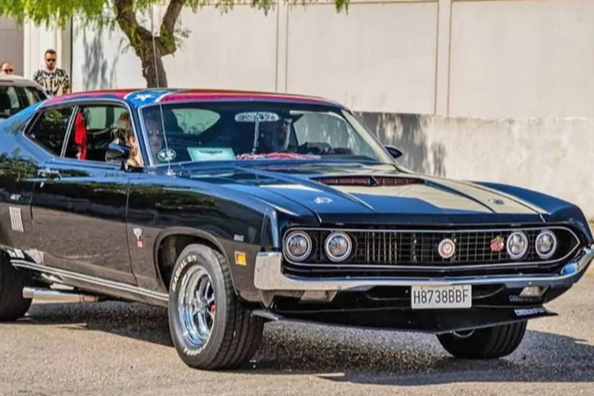 Auto clásico negro con detalles cromados circulando por una calle junto a una pared clara