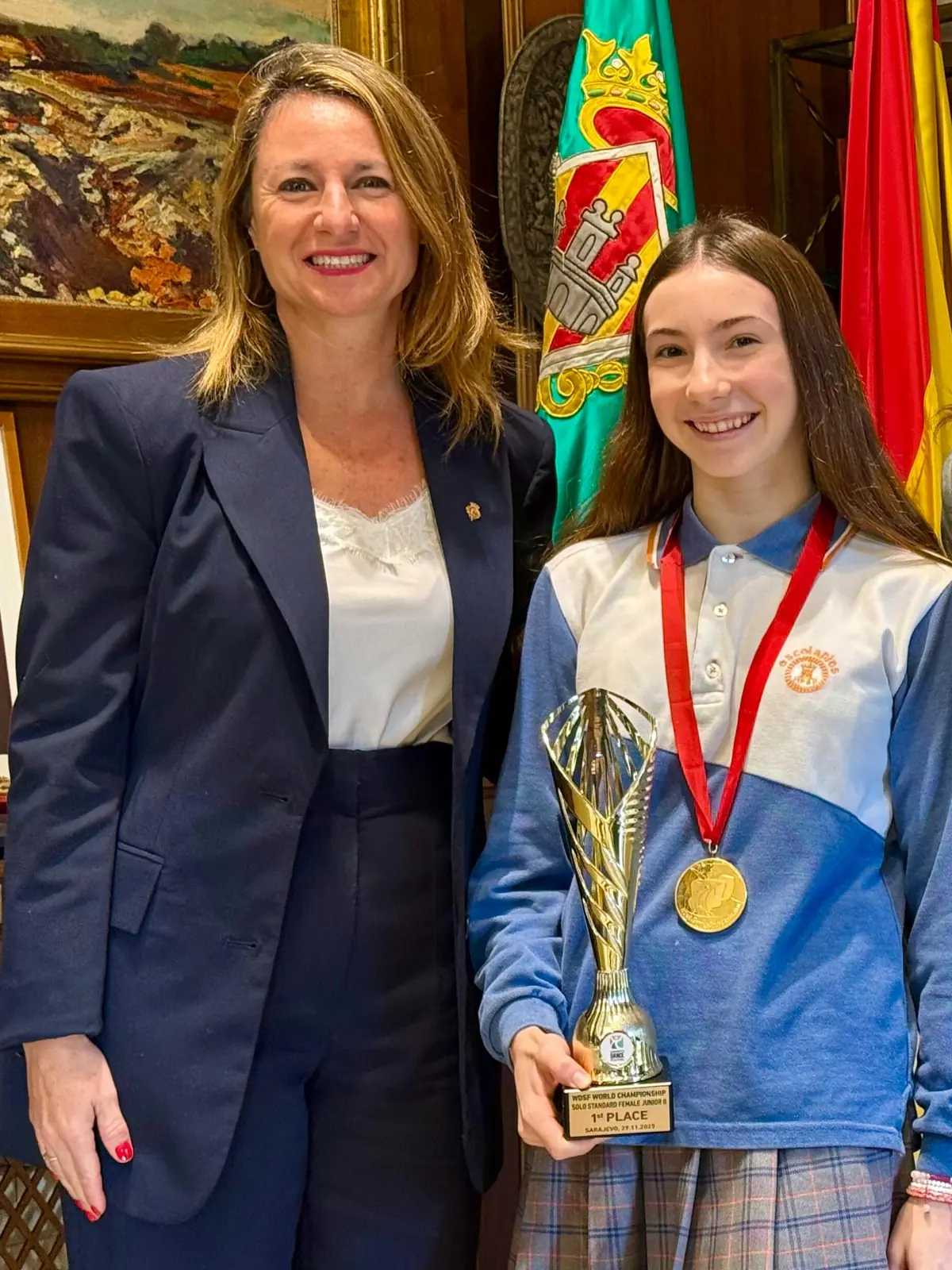 Dos mujeres sonríen mientras posan juntas, una de ellas sostiene un trofeo dorado y lleva una medalla, al fondo se ven banderas y un cuadro Dos mujeres sonríen mientras posan juntas, una de ellas sostiene un trofeo dorado y lleva una medalla, al fondo se ven banderas y un cuadro