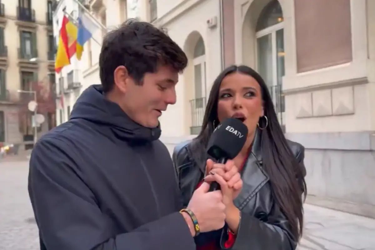 Un hombre y una mujer sonríen mientras sostienen juntos un micrófono de EDATV en una calle urbana con banderas al fondo