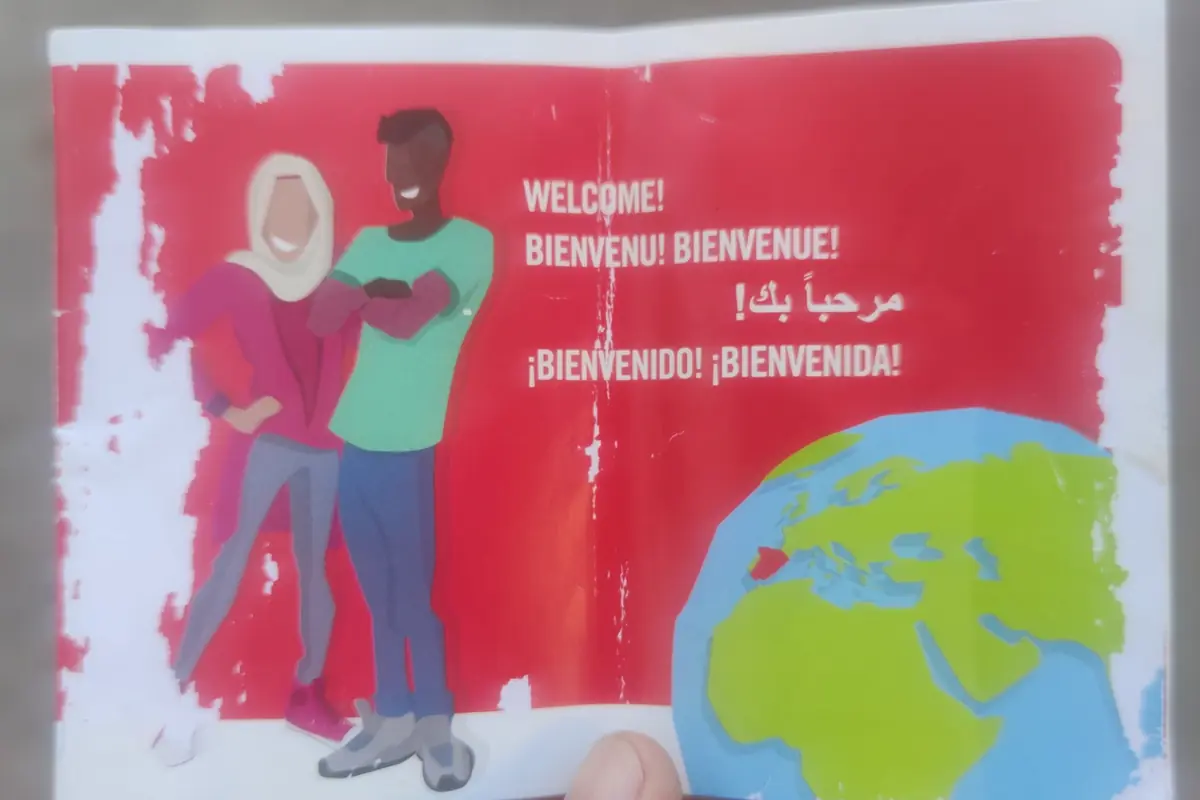 Folleto con fondo rojo que muestra a dos personas sonrientes junto a un globo terráqueo y varios mensajes de bienvenida en diferentes idiomas