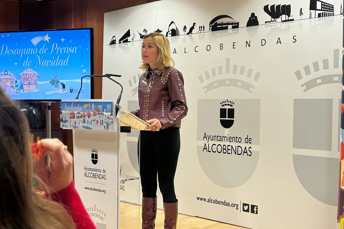 Una mujer rubia con chaqueta marrón y botas habla en un atril durante un evento navideño del Ayuntamiento de Alcobendas, con una pantalla al fondo que muestra el texto Desayuno de Prensa de Navidad y decoraciones festivas