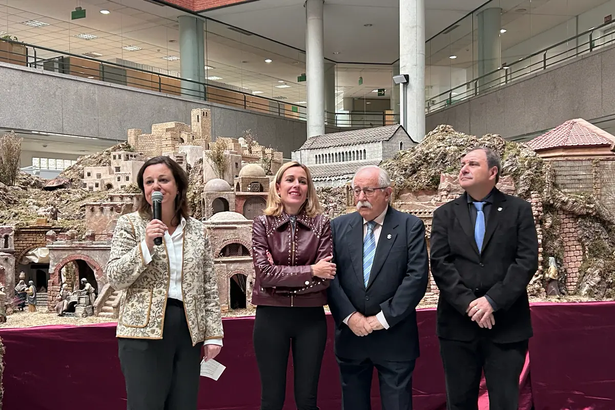 Cuatro personas posan frente a un gran belén navideño en el interior de un edificio moderno, una de ellas sostiene un micrófono mientras las otras tres observan sonrientes.