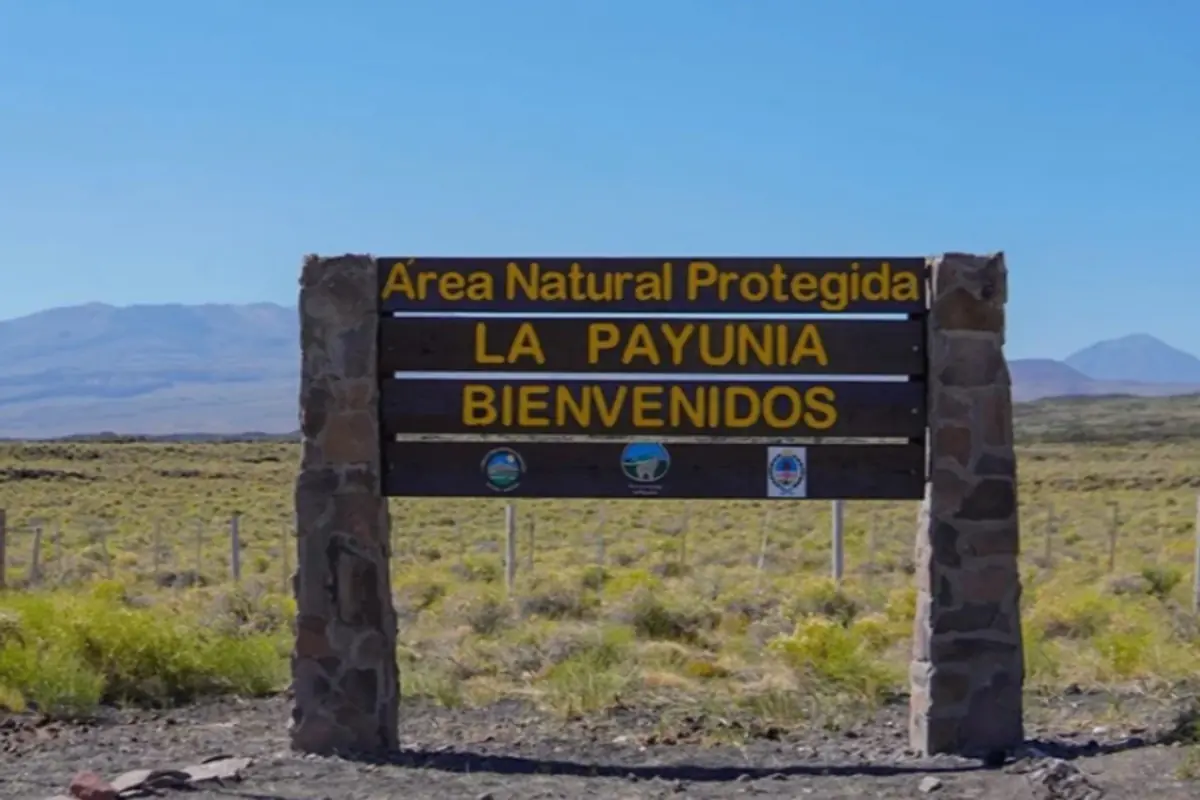Cartel de bienvenida al Área Natural Protegida La Payunia en un paisaje de campo abierto con montañas al fondo Cartel de bienvenida al Área Natural Protegida La Payunia en un paisaje de campo abierto con montañas al fondo