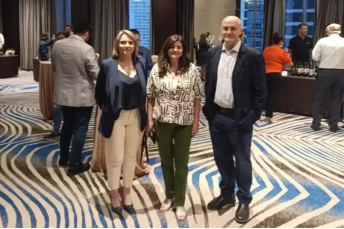 Tres personas posan para una foto en un salón elegante con alfombra de diseño geométrico y varias personas al fondo conversando y sirviéndose bebidas.