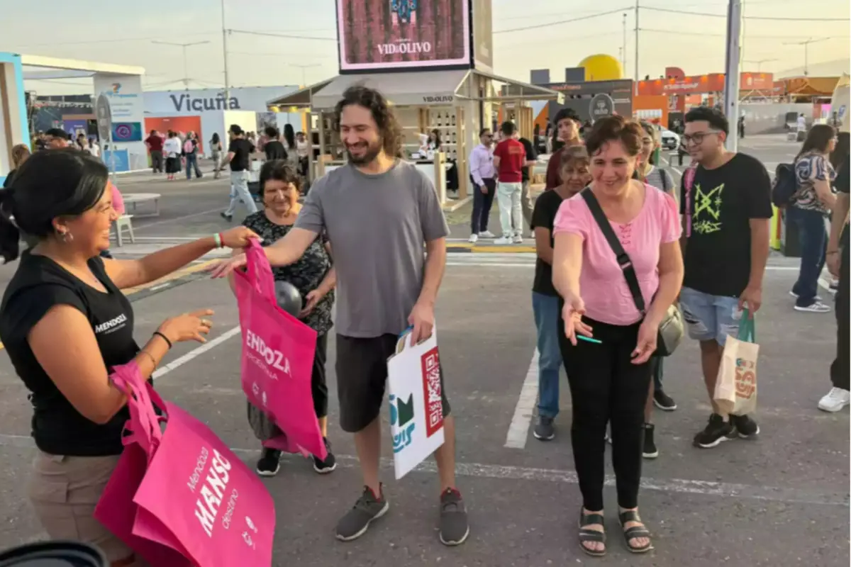 Una mujer entrega bolsas rosas promocionales a personas sonrientes en un evento al aire libre con varios puestos y gente caminando en el fondo