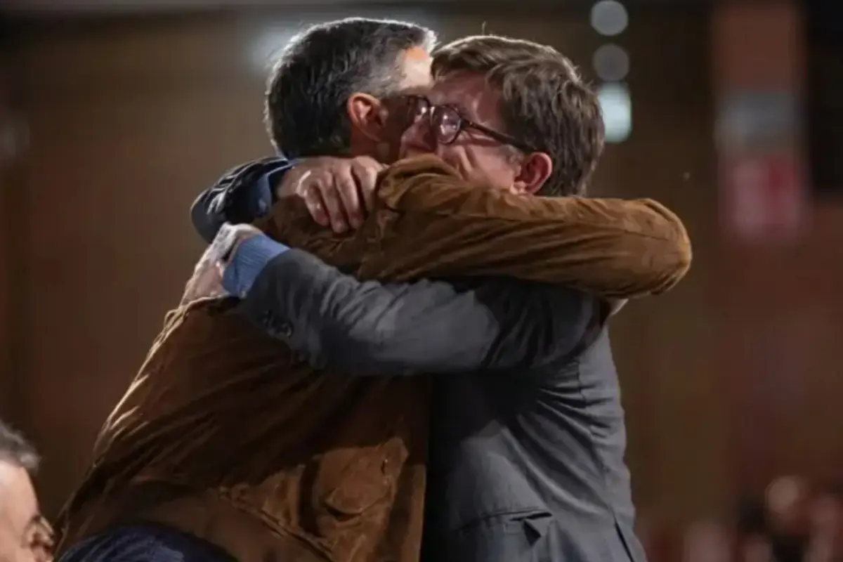 Dos hombres se abrazan con fuerza mostrando una expresión de emoción y alegría en un ambiente interior.