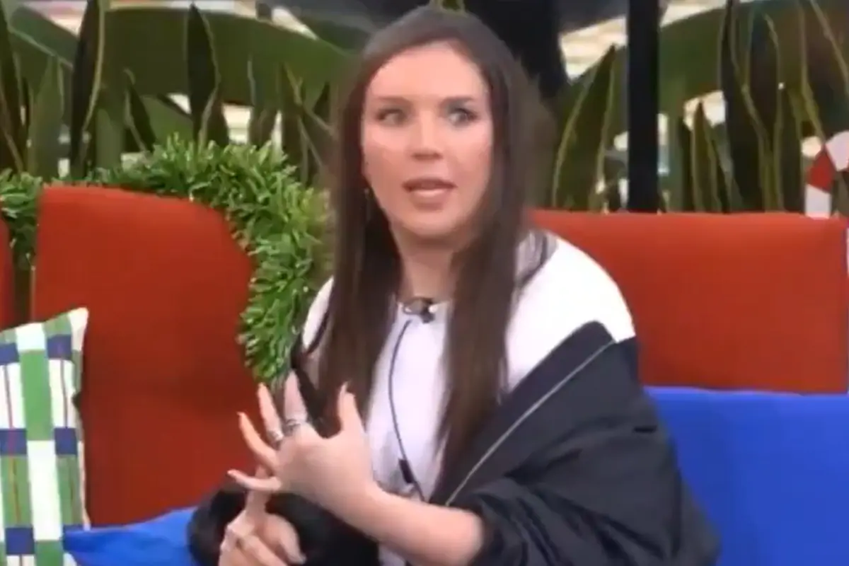 Mujer de cabello largo y lacio con expresión de sorpresa sentada en un sillón rojo con cojines y plantas al fondo