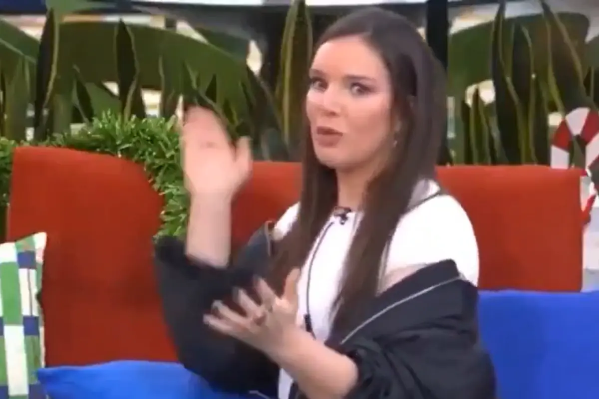 Mujer con expresión de sorpresa levantando la mano mientras está sentada en un sillón rojo con cojines y plantas al fondo