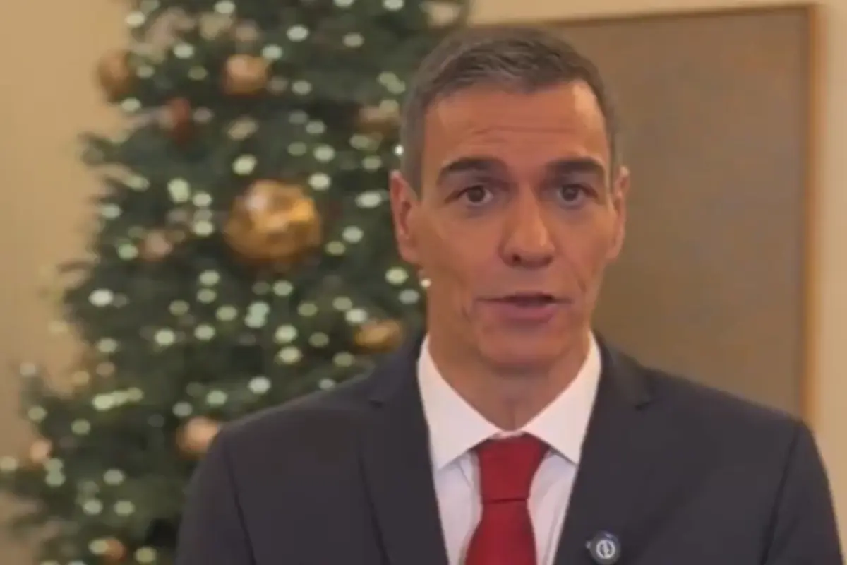 Hombre con traje oscuro y corbata roja hablando frente a un árbol de Navidad decorado con luces y bolas doradas