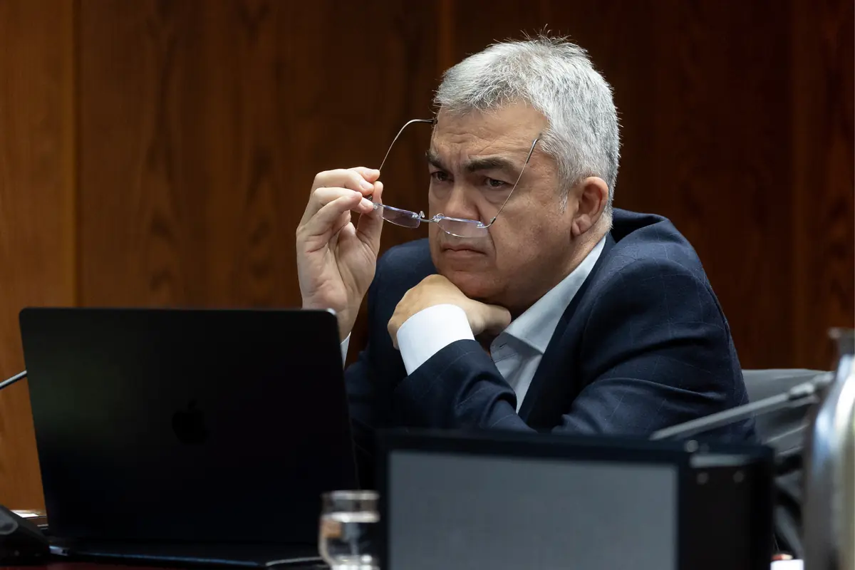 Hombre de mediana edad con traje oscuro se quita las gafas mientras mira pensativo la pantalla de un portátil en una sala de reuniones de madera Hombre de mediana edad con traje oscuro se quita las gafas mientras mira pensativo la pantalla de un portátil en una sala de reuniones de madera