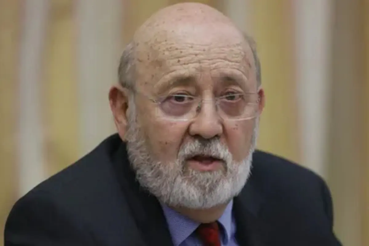 Hombre mayor con barba y gafas usando saco oscuro y corbata roja