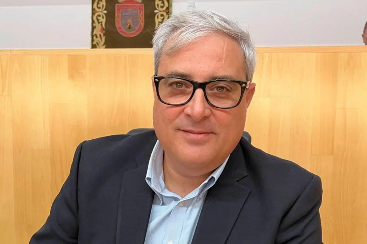 Hombre de cabello canoso y gafas negras sentado frente a un fondo de madera clara con un escudo en la pared Hombre de cabello canoso y gafas negras sentado frente a un fondo de madera clara con un escudo en la pared