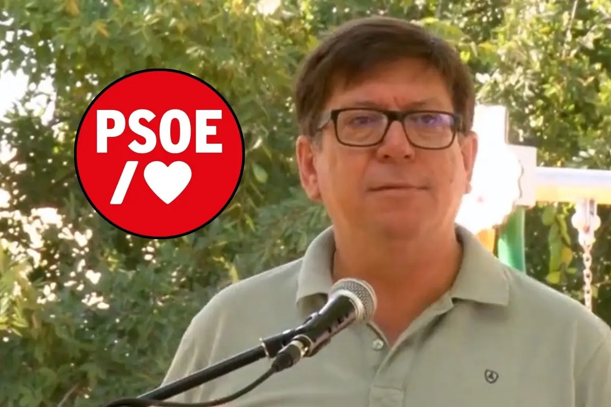 Hombre con gafas hablando ante un micrófono al aire libre junto a un gran círculo rojo con el logotipo del PSOE y un corazón blanco