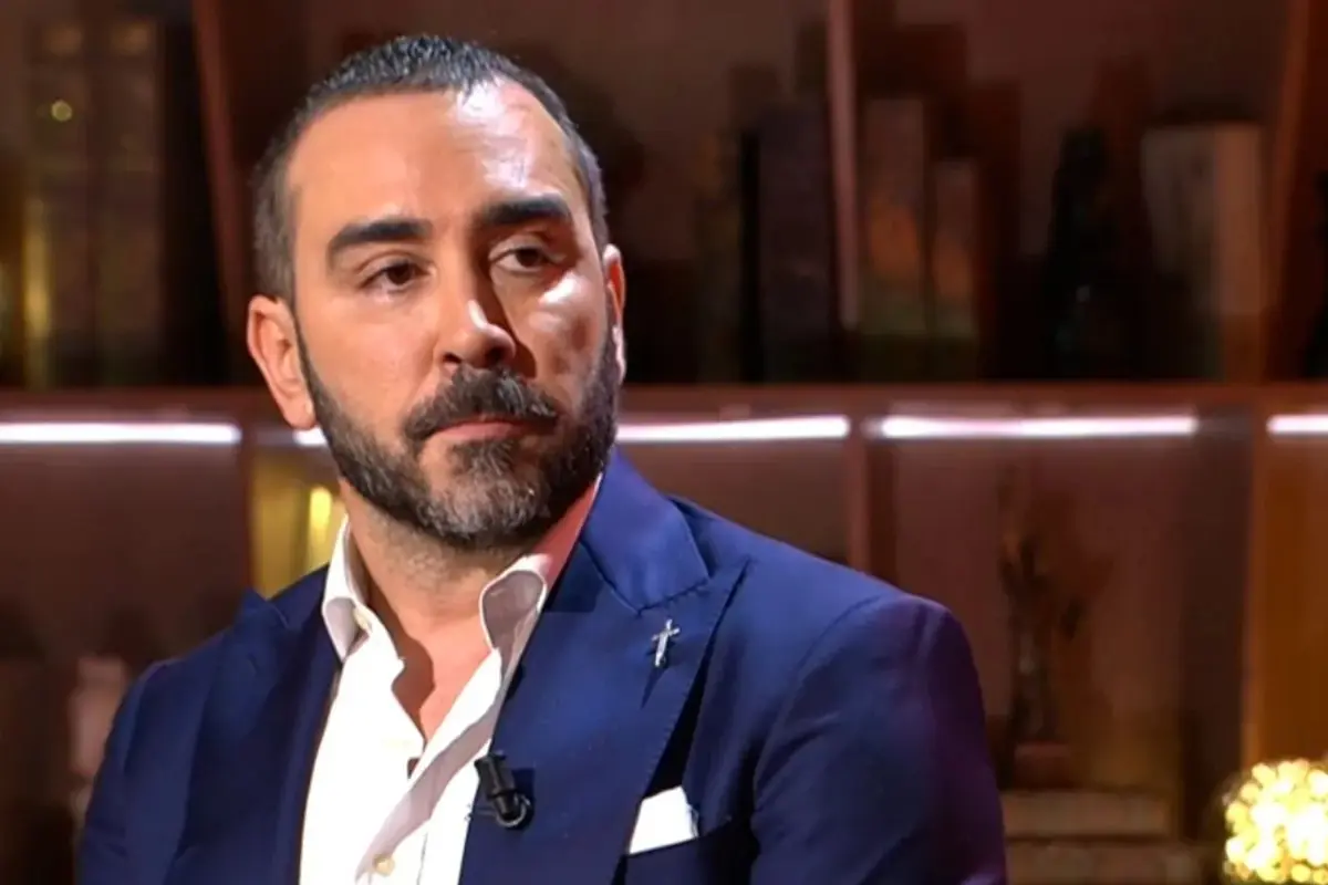 Hombre de mediana edad con barba y traje azul marino sentado en un plató de televisión con expresión seria Hombre de mediana edad con barba y traje azul marino sentado en un plató de televisión con expresión seria