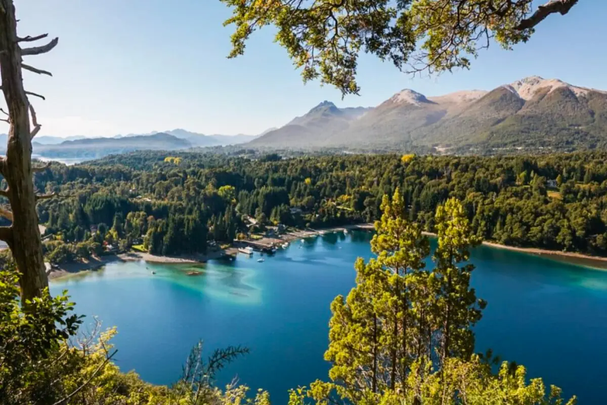 Vista panorámica de un lago de aguas cristalinas rodeado de bosques verdes y montañas bajo un cielo despejado