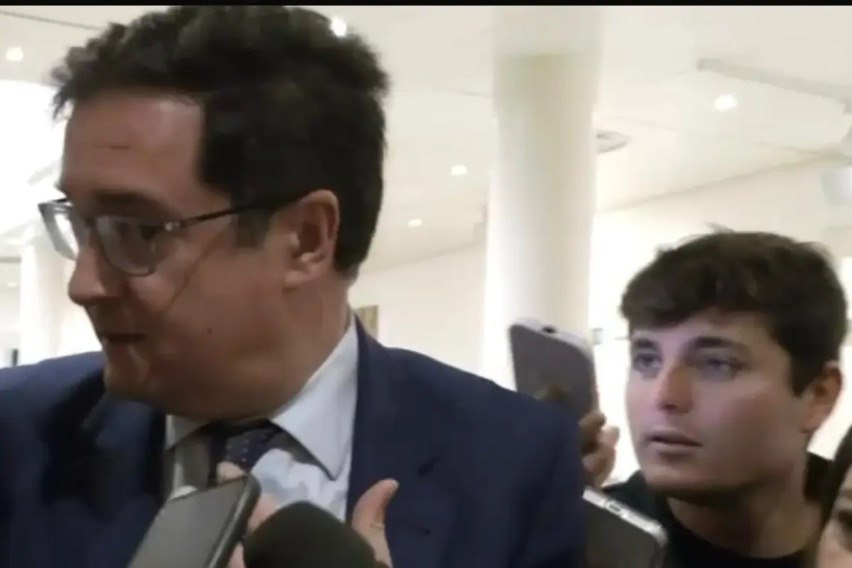 Hombre con traje azul respondiendo a periodistas mientras otro joven lo graba con el móvil en un pasillo iluminado Hombre con traje azul respondiendo a periodistas mientras otro joven lo graba con el móvil en un pasillo iluminado