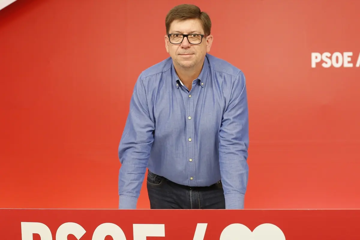 Hombre con gafas y camisa azul apoyado sobre un atril rojo con el logotipo del PSOE delante de un fondo rojo Hombre con gafas y camisa azul apoyado sobre un atril rojo con el logotipo del PSOE delante de un fondo rojo
