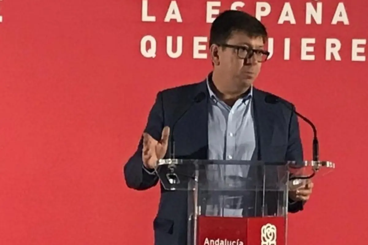 Hombre con gafas hablando en un atril transparente durante un acto político del PSOE en Andalucía con fondo rojo y texto parcial La España que quieres Hombre con gafas hablando en un atril transparente durante un acto político del PSOE en Andalucía con fondo rojo y texto parcial La España que quieres