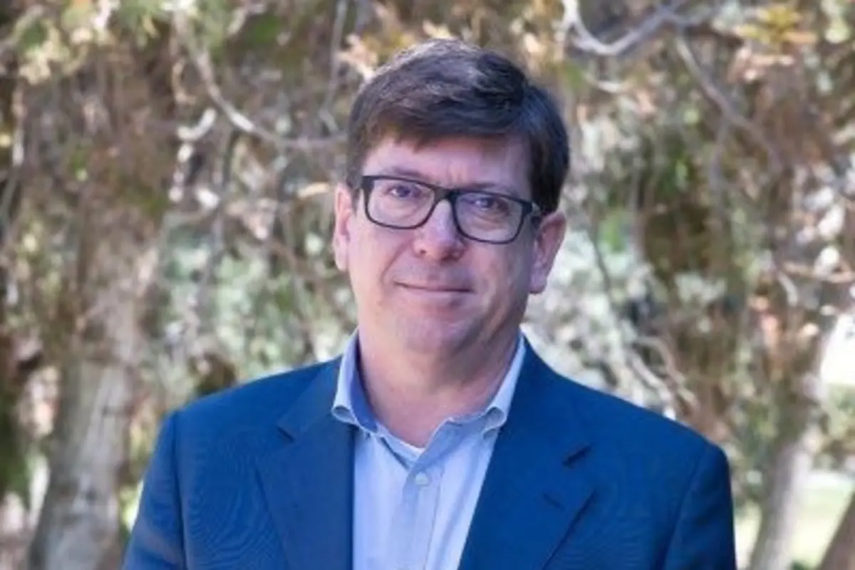 Hombre de cabello castaño y gafas con saco azul posando al aire libre frente a árboles desenfocados
