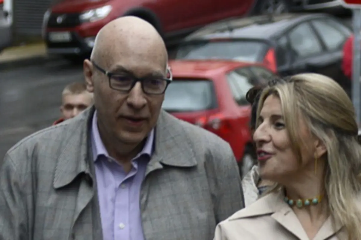 Hombre calvo con gafas y mujer rubia conversando mientras caminan por una calle con coches aparcados de fondo