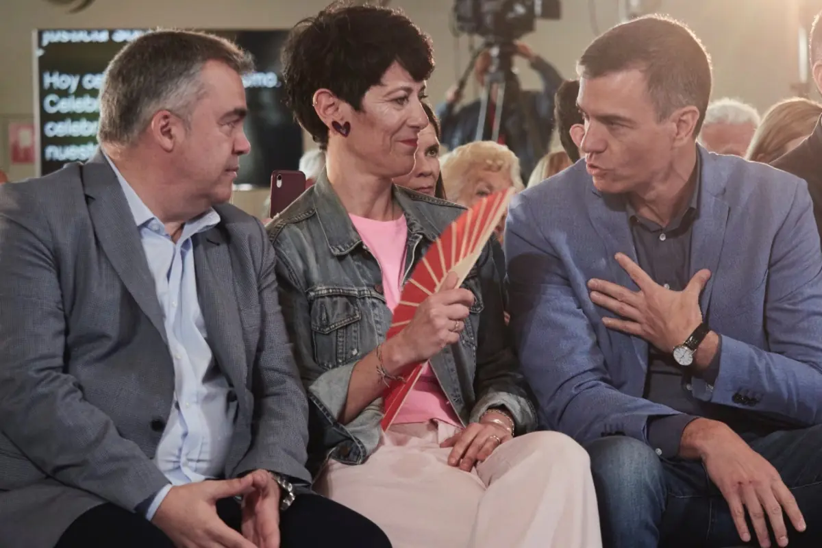 Tres personas sentadas en primera fila conversan en un acto público mientras la mujer del centro sostiene un abanico rojo y mira al hombre de la derecha que gesticula con la mano en el pecho