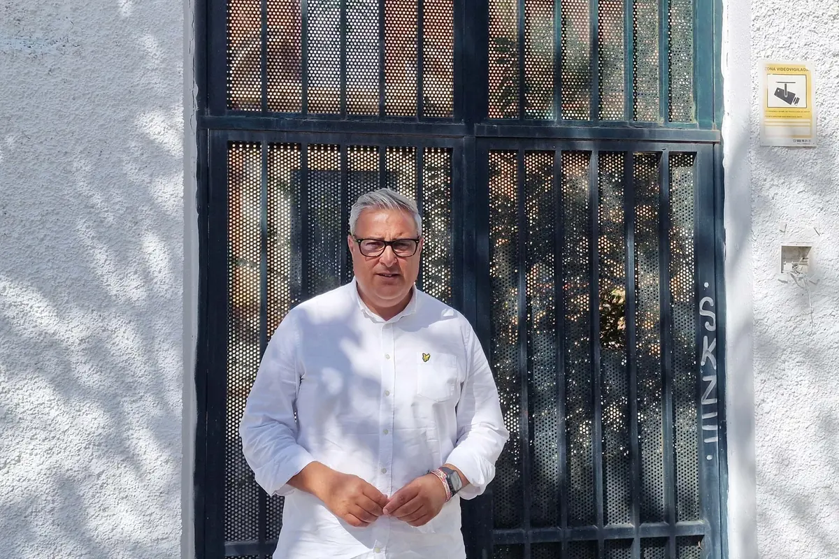 Hombre de cabello canoso y gafas con camisa blanca de pie frente a una puerta metálica negra