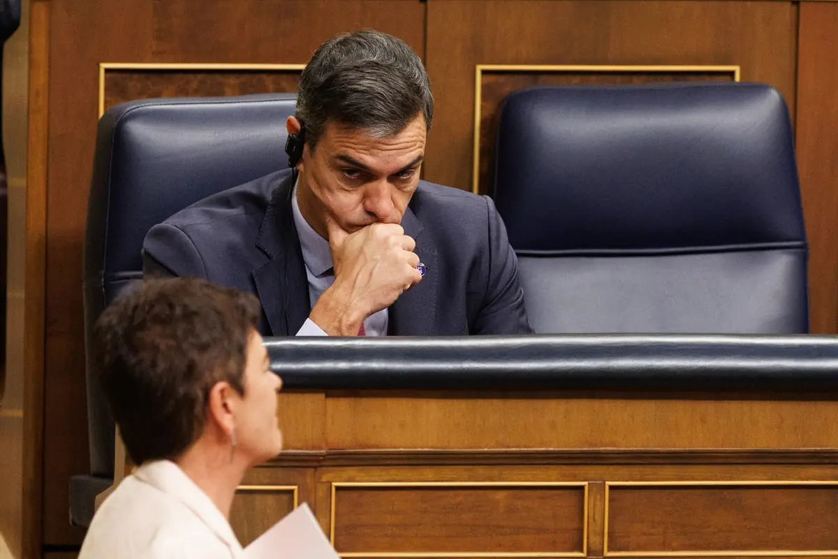 Hombre con expresión seria y pensativa sentado en un escaño de un parlamento mientras una mujer habla en primer plano