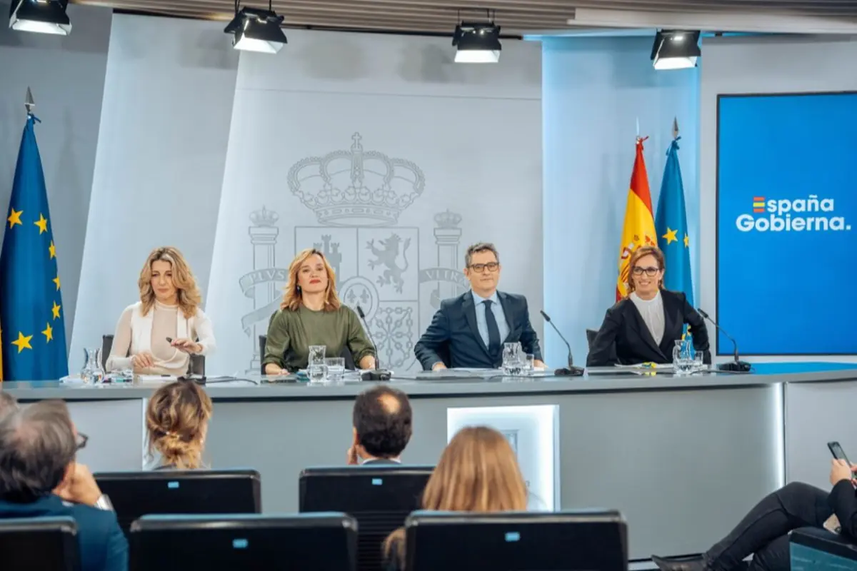Cuatro personas sentadas en una mesa durante una rueda de prensa oficial con banderas de la Unión Europea y de España al fondo y una pantalla que muestra el texto España Gobierna