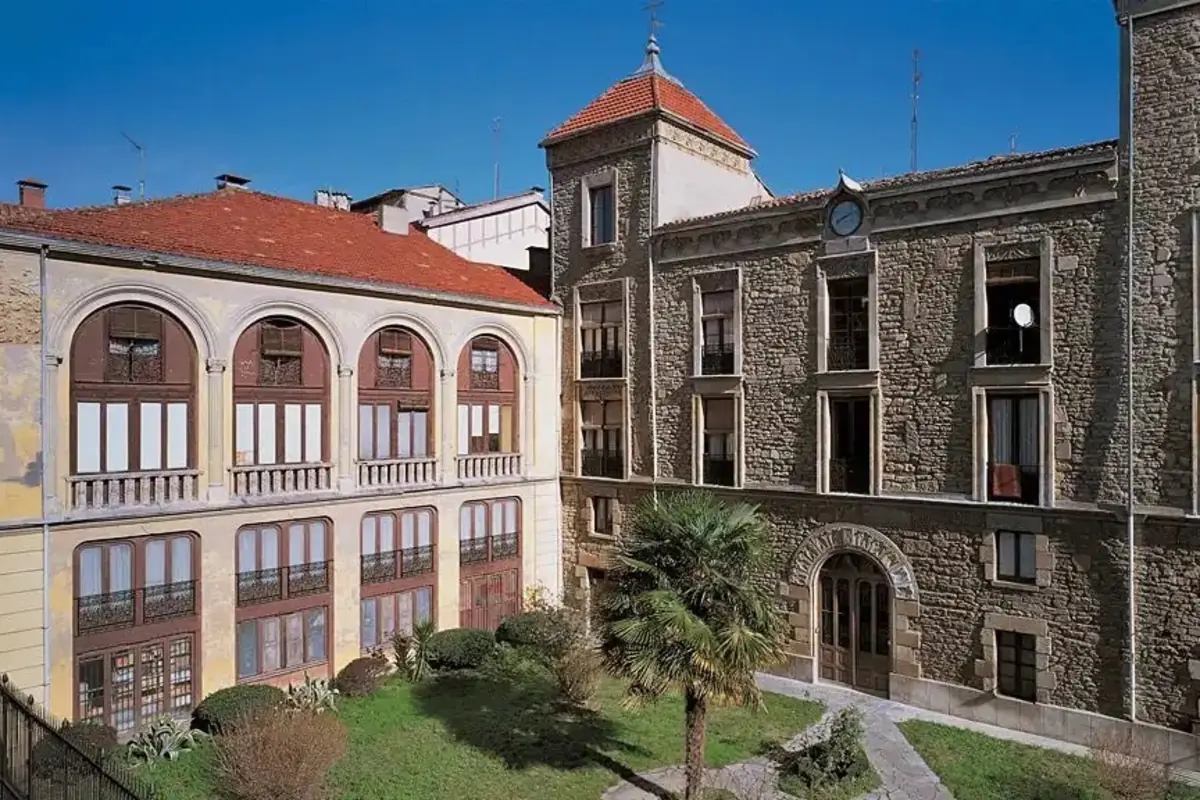 Edificio antiguo de piedra con techo rojo y ventanas grandes rodeando un pequeño jardín con palmeras y arbustos bajo un cielo despejado